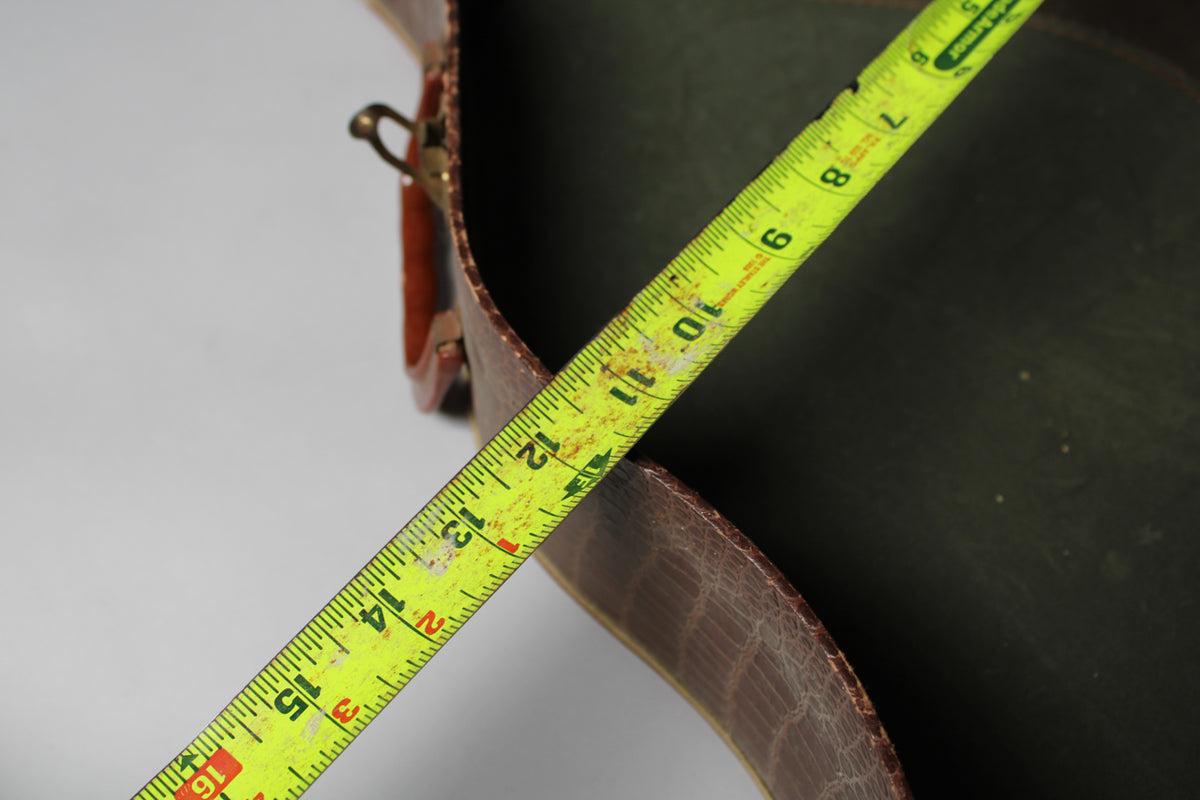 A yellow measuring tape spans the open, dark green interior of the brown snakeskin-patterned guitar case.