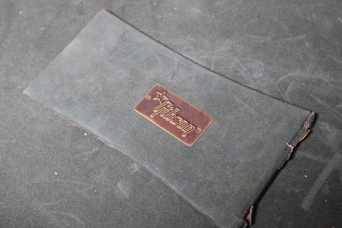 A dark green, rectangular paper tag with a raised copper-colored Gibson logo, showing wear and a torn edge.