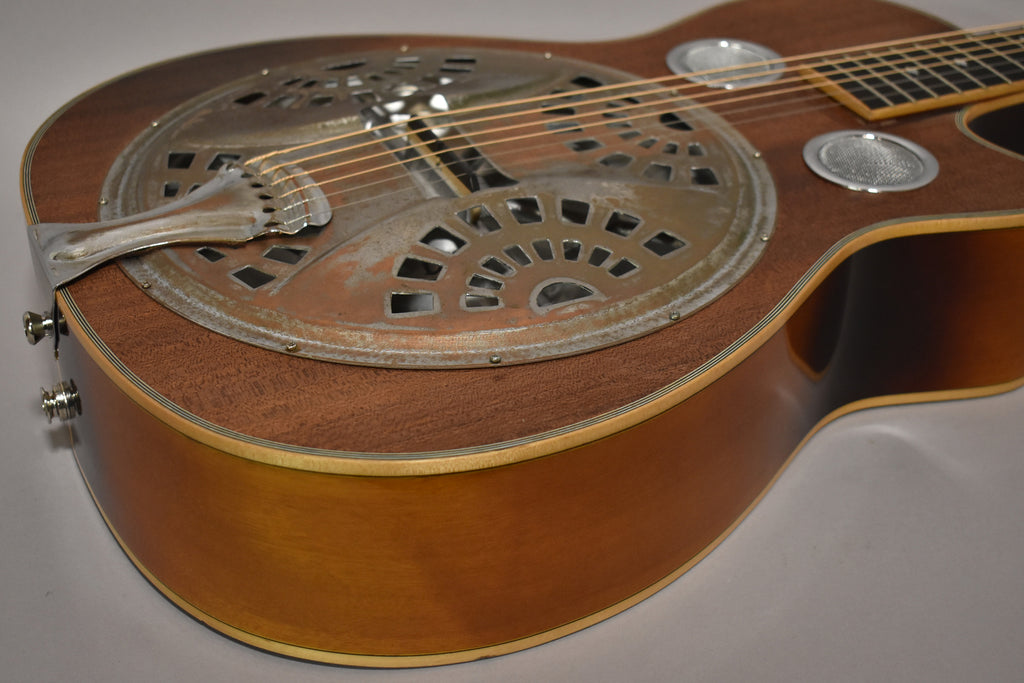 The resonator guitar's body, angled from the lower bout, shows a worn, tarnished metal resonator cover and a warm brown finish.