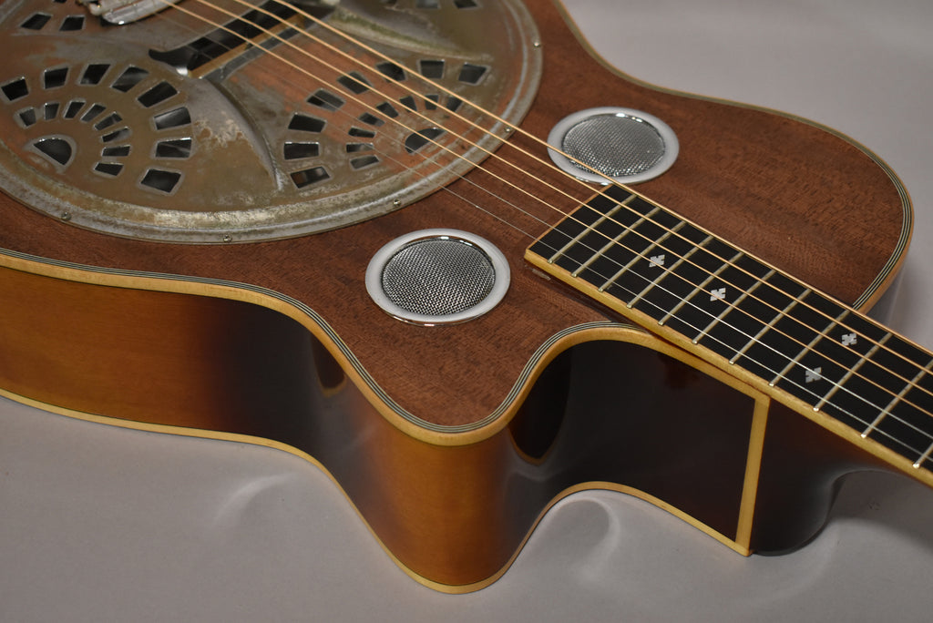 Close-up of the resonator guitar's body, showing the distressed metal resonator, two round soundholes, and the dark wood finish.