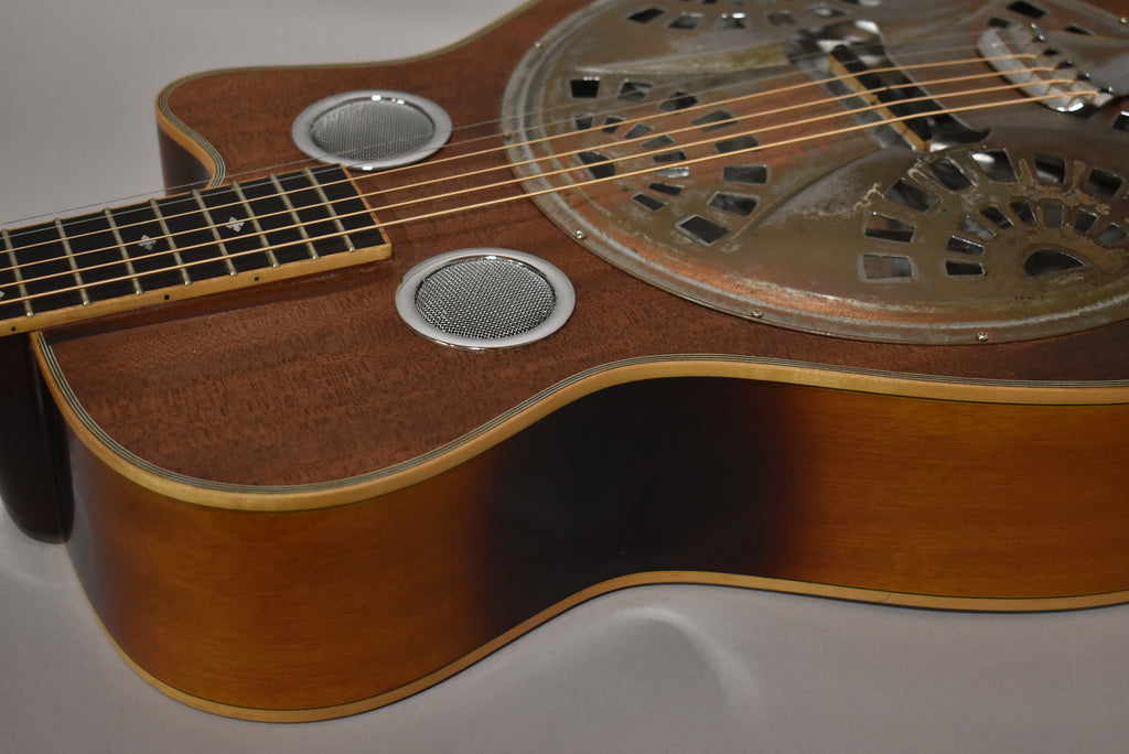 Close-up of the resonator guitar's body, showing its dark wood grain, two round chrome sound screens, and the tarnished metal resonator.
