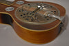 The resonator guitar's body, with a dark brown top and lighter sides, shows a tarnished, ornate metal resonator cover.