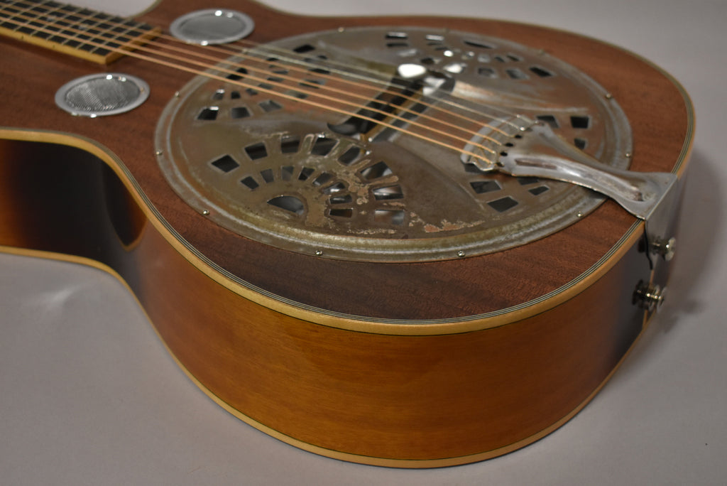 The resonator guitar's body, with a dark brown top and lighter sides, shows a tarnished, ornate metal resonator cover.