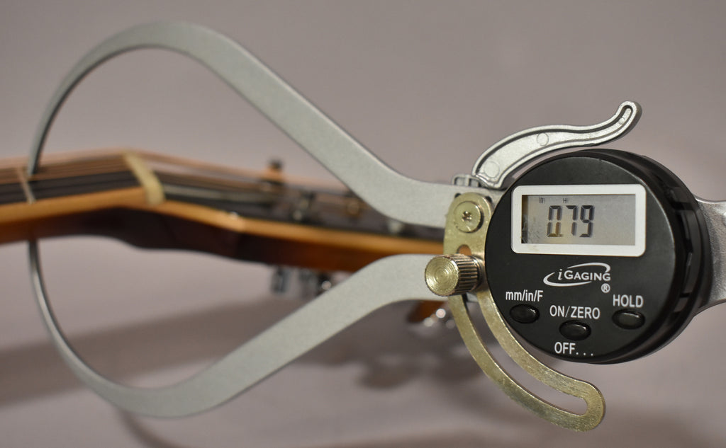 A digital caliper measures the string height at the nut of the guitar, displaying "0.79" on its screen.