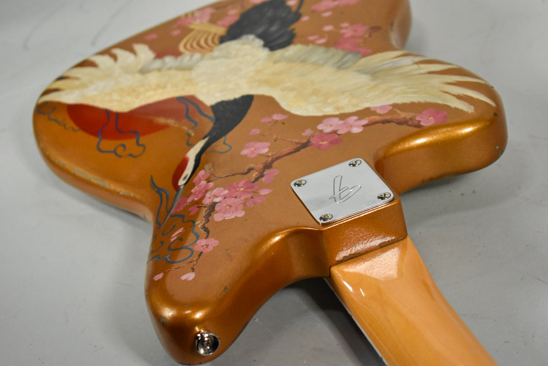 The guitar's metallic orange body, adorned with a hand-painted crane and cherry blossoms, shows light wear near the neck plate.