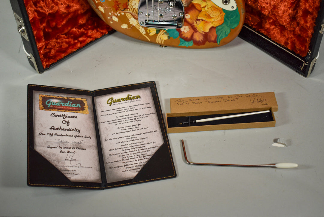 Top-down view of a black certificate folder, a brown box with a tremolo arm, and a guitar body with floral art.