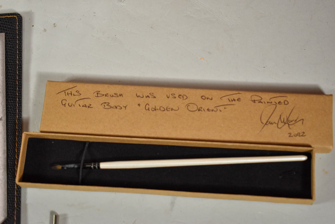 An open cardboard box containing a white-handled paintbrush on black foam, with handwritten text and a signature on the box lid.