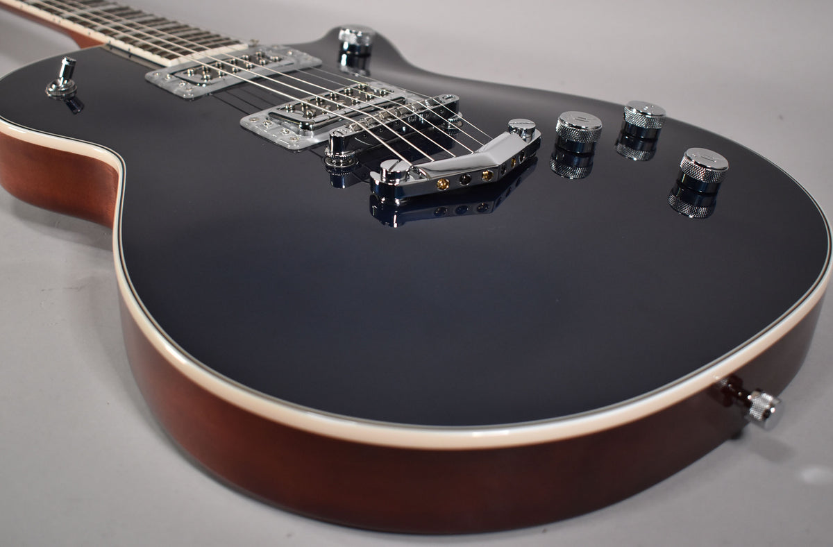 The guitar's dark grey top and brown sides are visible from a low angle, showing chrome hardware and four knurled control knobs.