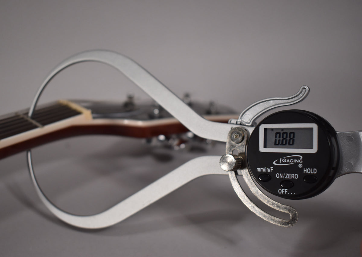 A digital caliper measures the guitar's nut width, displaying "0.88" on its screen, with the headstock blurred in the background.