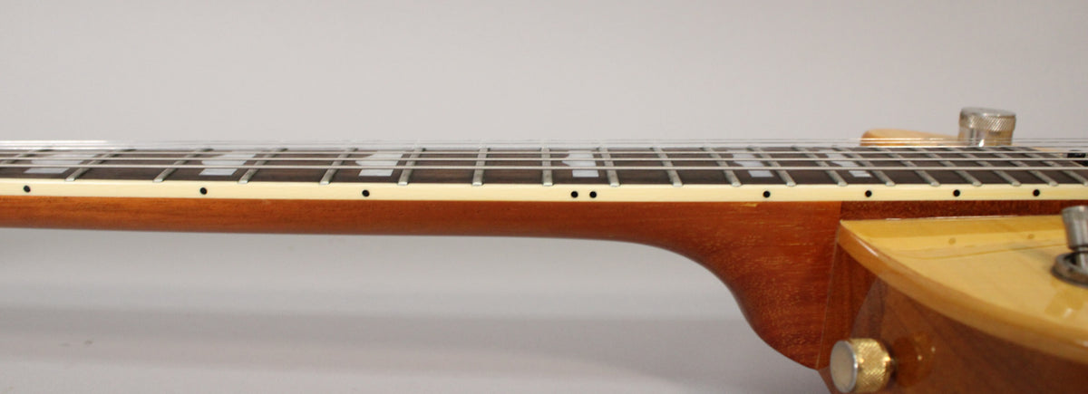 The guitar's neck, with a dark brown finish, is shown from the side, revealing cream binding and block inlays on the fretboard.