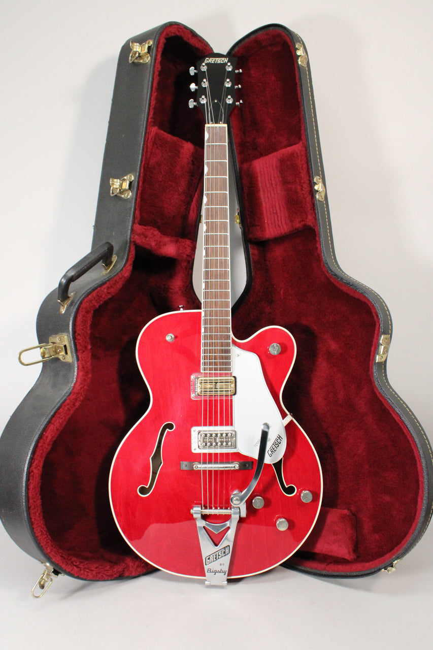 Full-length view of a glossy red hollow-body electric guitar with a white pickguard and Bigsby tremolo, standing in its open, red-lined hard case.