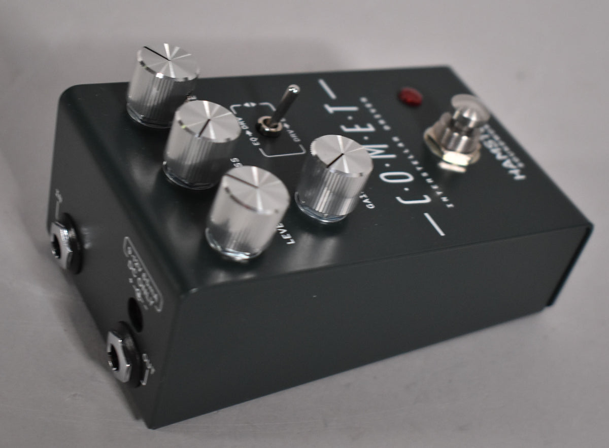 A dark gray pedal is angled from the left, showing its top with four silver knobs, a toggle switch, a red LED, and a footswitch.