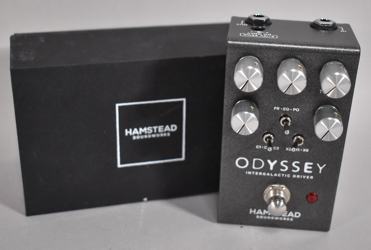 A dark gray guitar pedal with five silver knobs, three toggle switches, a footswitch, and a red indicator light, next to a black box.