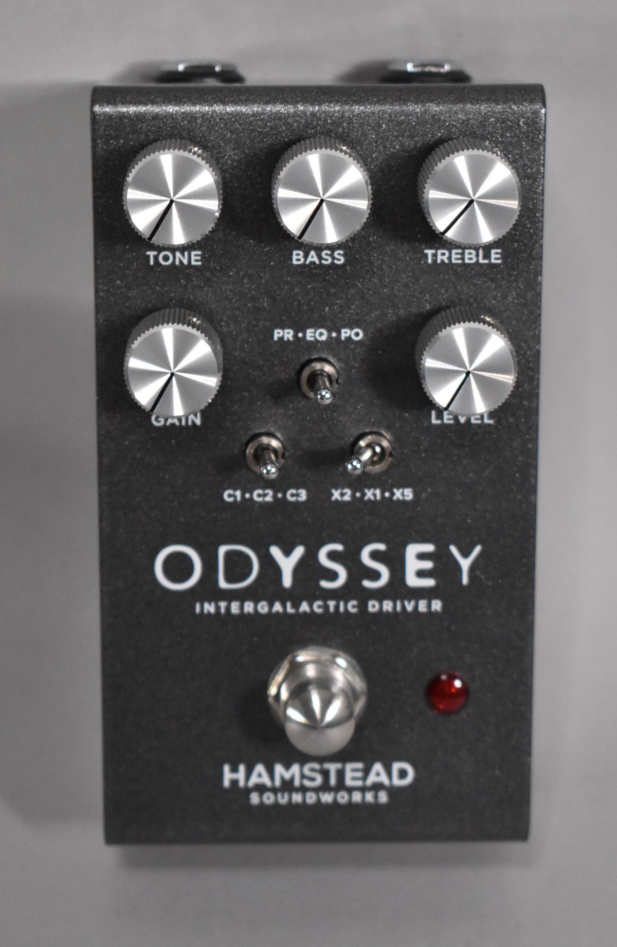 A top-down view of a dark gray textured pedal with five silver knobs, three mini-toggle switches, a footswitch, and a red LED.