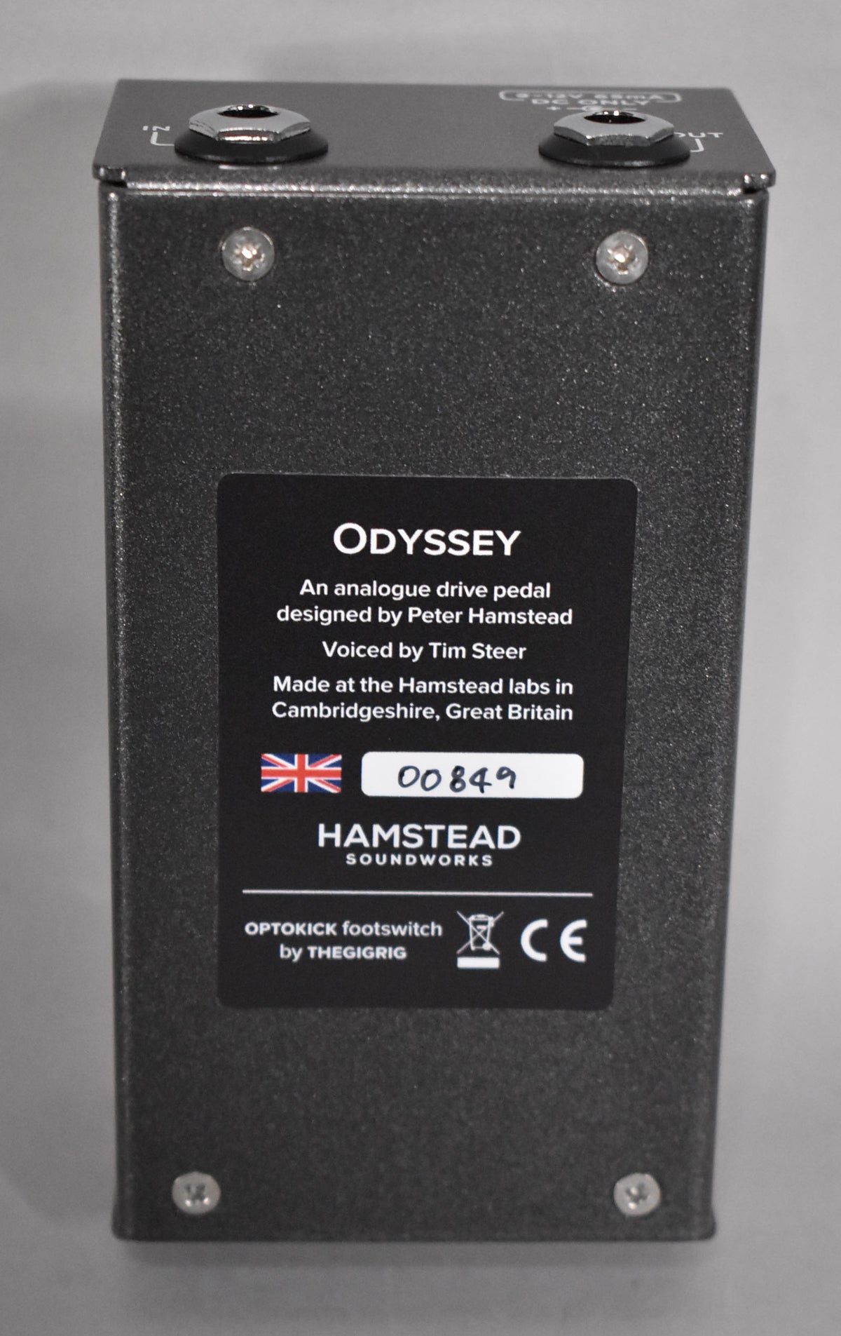 The dark gray textured metal pedal is viewed from the back, showing the input and output jacks and a black informational sticker.