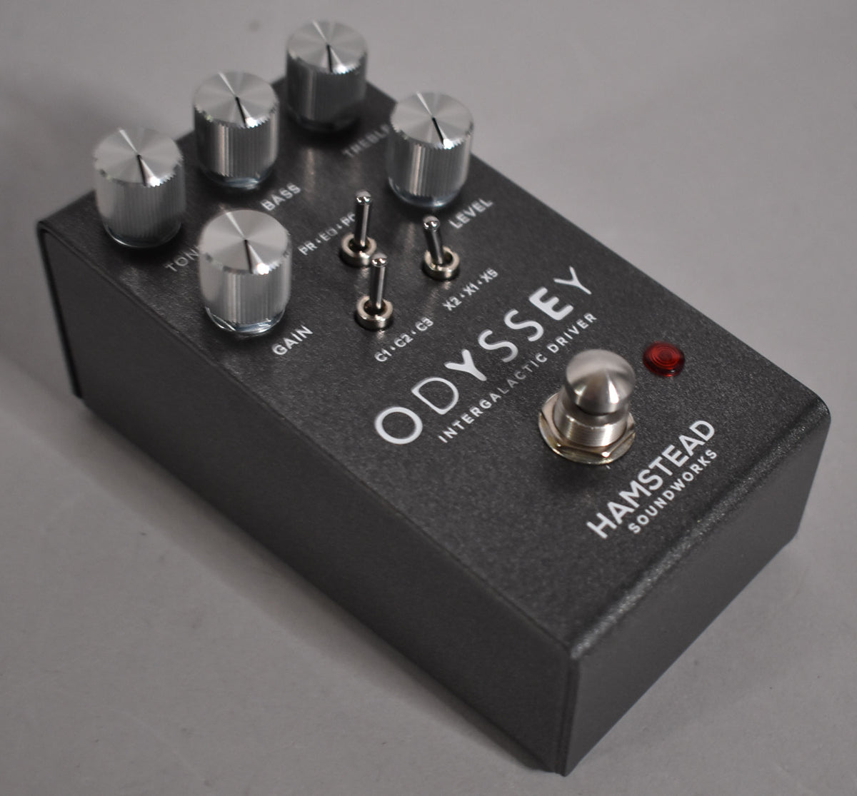 A dark gray textured overdrive pedal, viewed from above, with five silver knurled knobs, three silver toggle switches, and a silver footswitch.