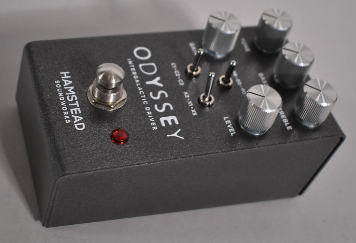 A dark gray overdrive pedal, angled from the left, shows a footswitch, red LED, three toggle switches, and five silver knurled knobs.