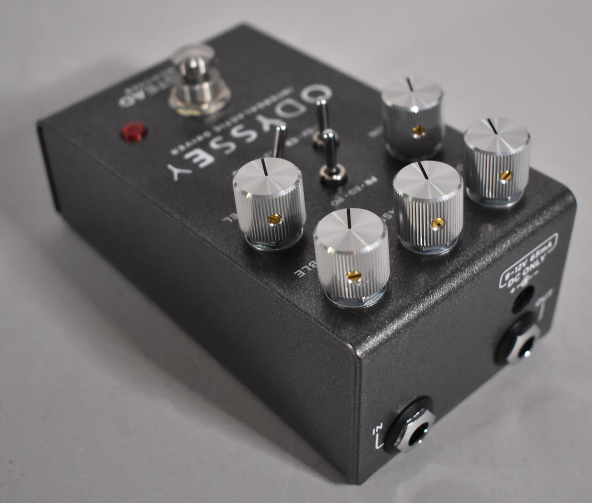 A dark gray overdrive pedal, viewed from the top right, showing five silver knurled knobs, two toggle switches, and a footswitch.