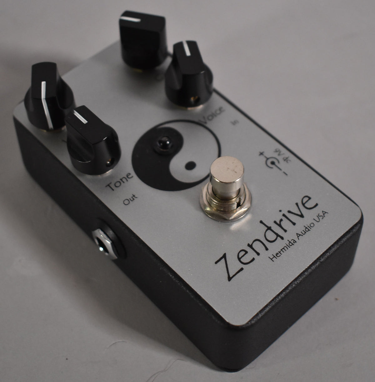 A silver guitar pedal with a black Yin-Yang symbol, four black control knobs, and a silver footswitch, viewed from the top-right.