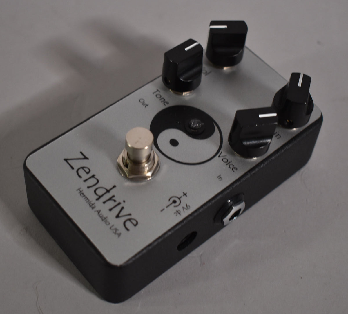 A silver-faced pedal with a black base, four black control knobs, a silver footswitch, and a yin-yang symbol.