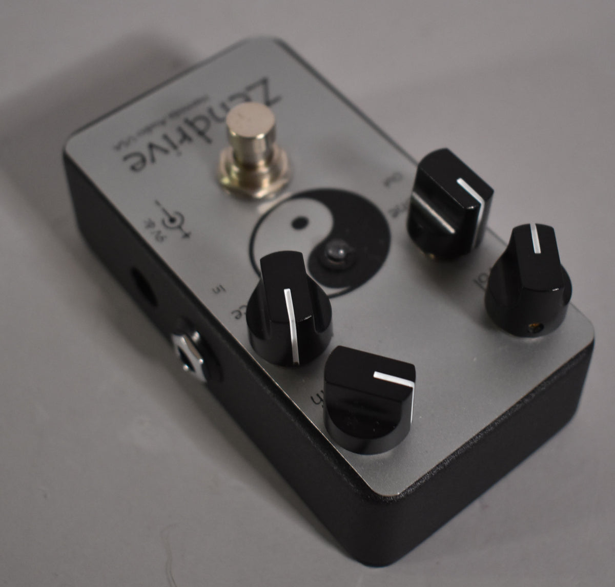 Angled view of a silver pedal with a black base, a yin-yang symbol, a silver footswitch, and four black knobs with white indicator lines.