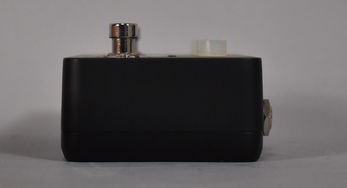 The black pedal is viewed from the side, showing a silver footswitch and a translucent white knob on top, with a silver jack on the right.