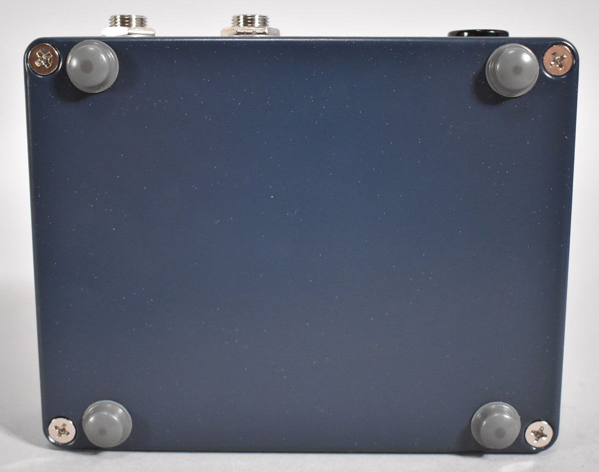 The dark blue pedal is viewed from the bottom, showing four grey rubber feet and four silver screws on a matte finish.