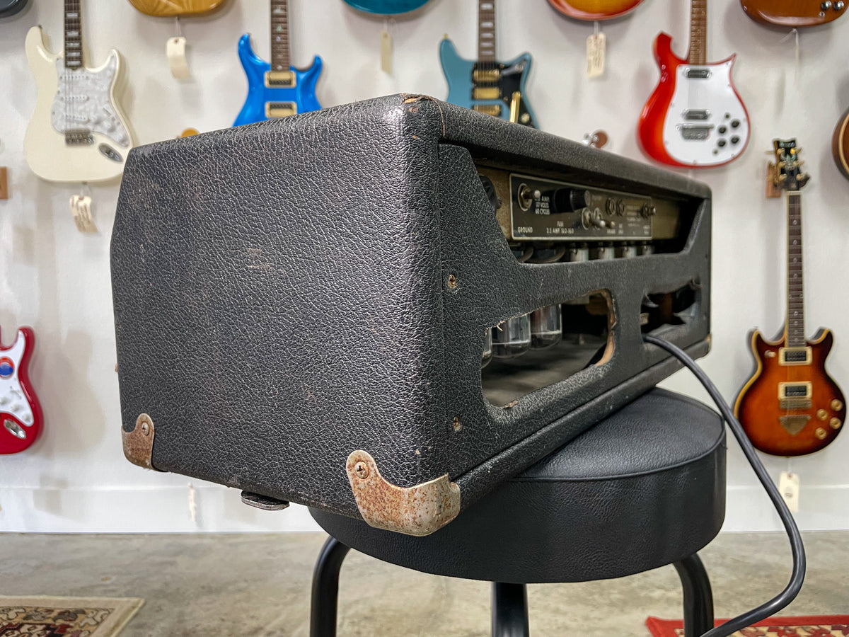 Black textured amp head, seen from the left rear, showing rusted corner protectors, exposed tubes, and rear panel controls.