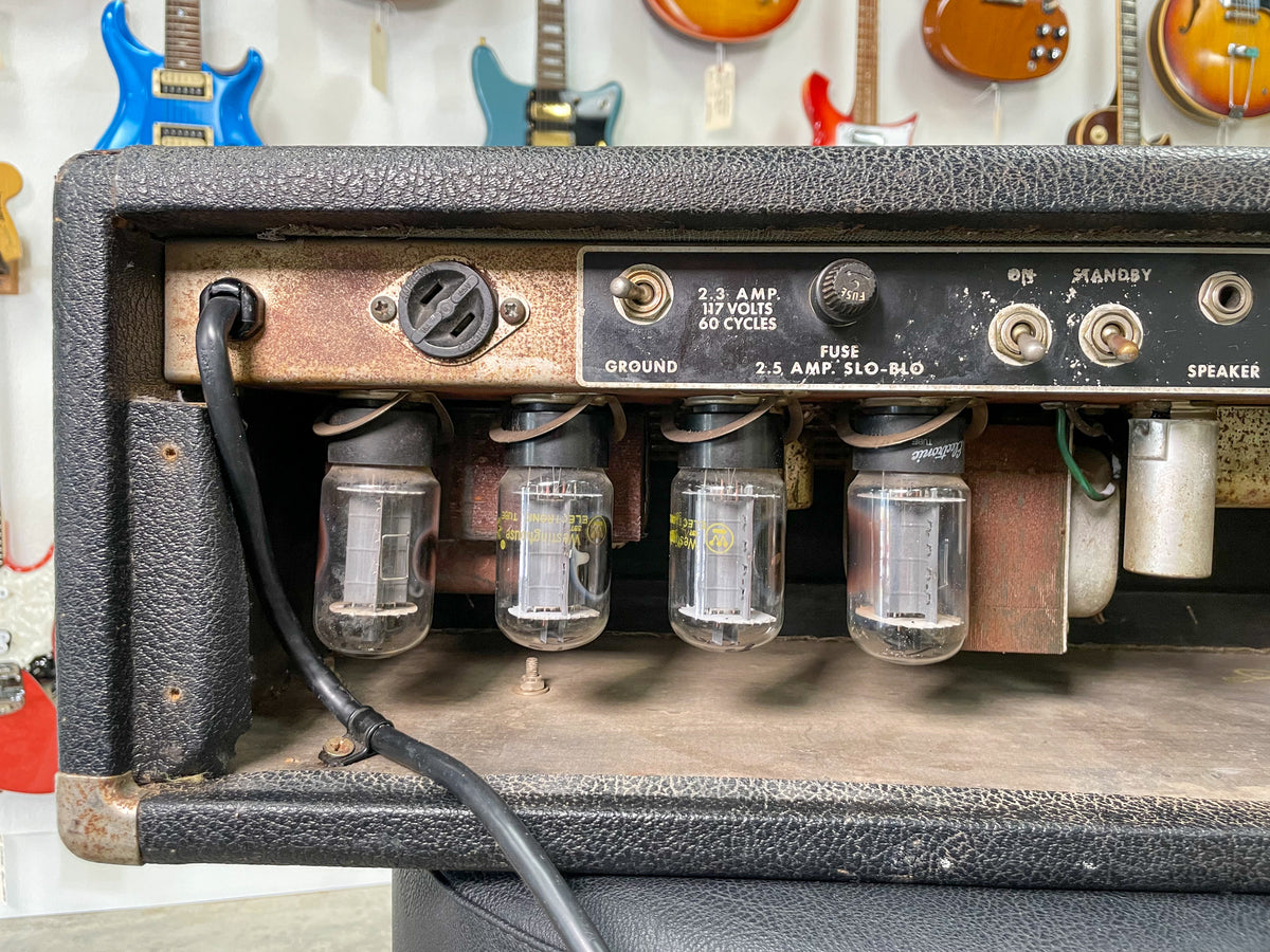 Rear view of the black tolex amp head, showing four glowing vacuum tubes, power cable, and the rusty back panel with controls.