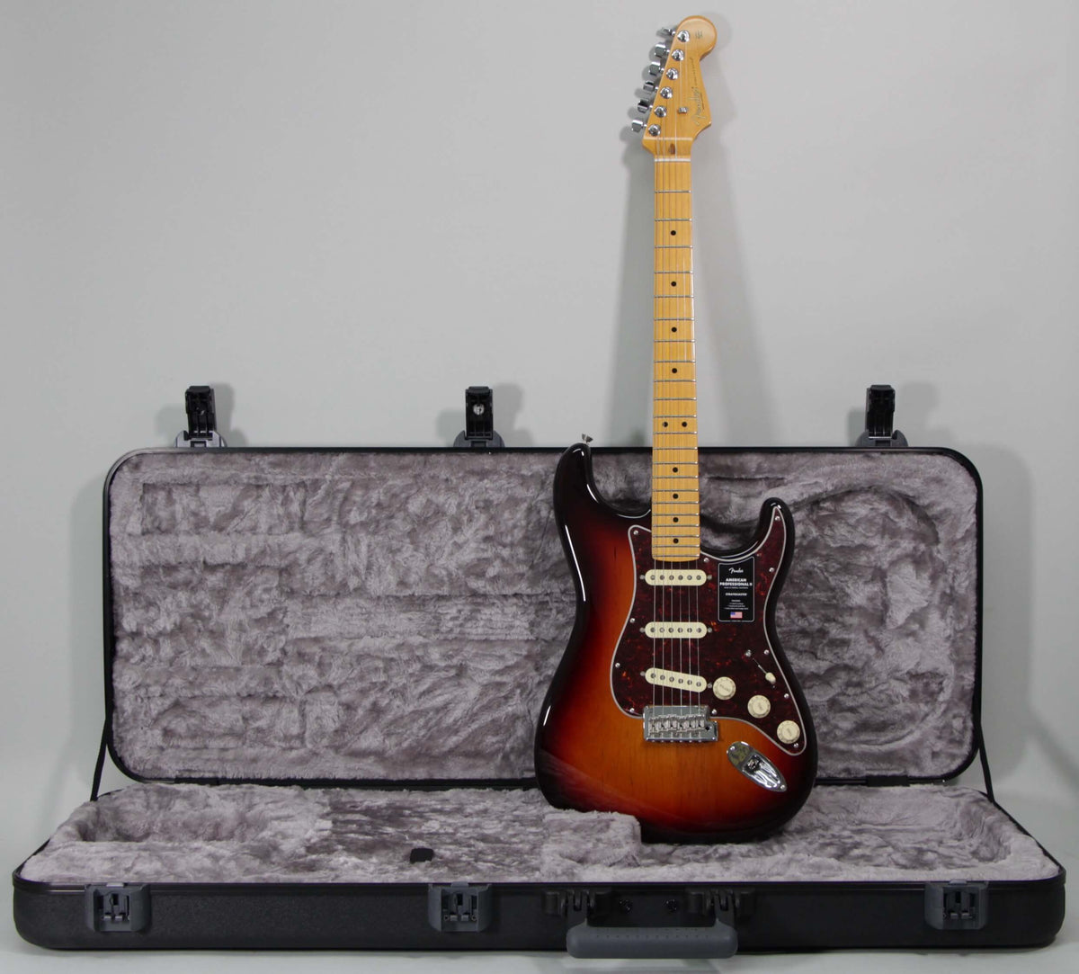 Full-length view of a sunburst Stratocaster with a maple neck, standing upright in front of its open, grey-lined hard case.