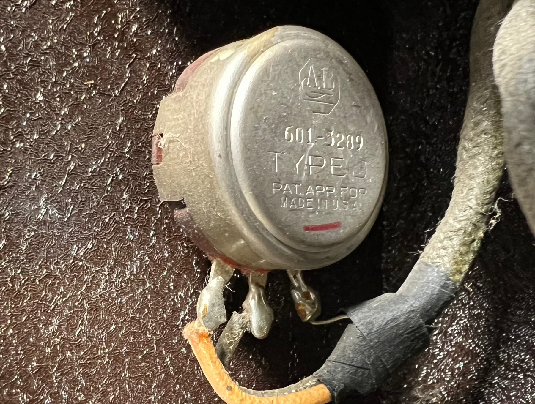 Close-up of a silver, circular potentiometer with three soldered terminals and an orange wire, against a dark, textured background.