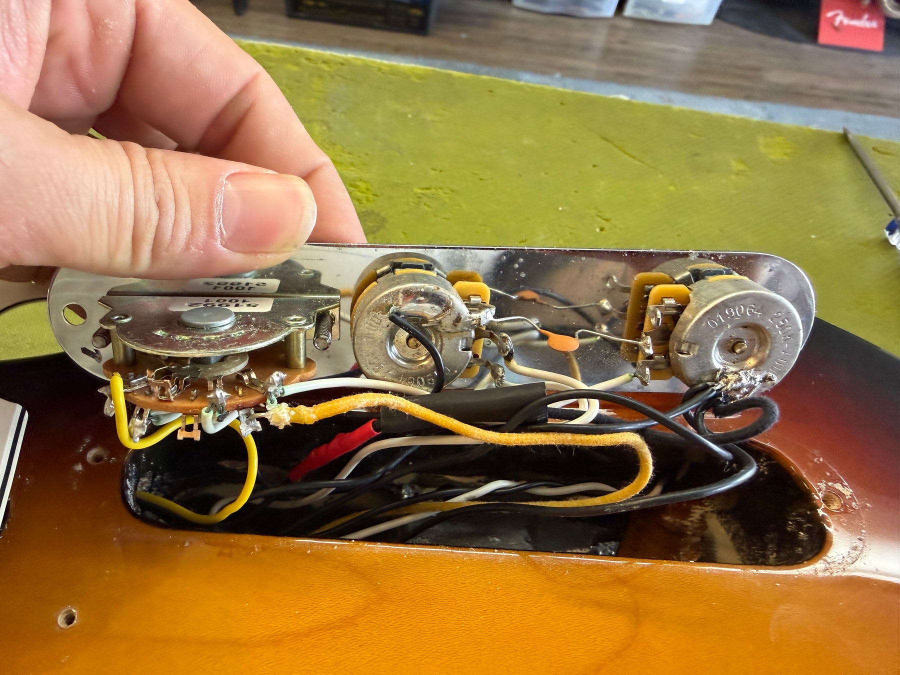 A hand holds the chrome control plate, revealing the guitar's wiring, pots, and switch over the sunburst body.