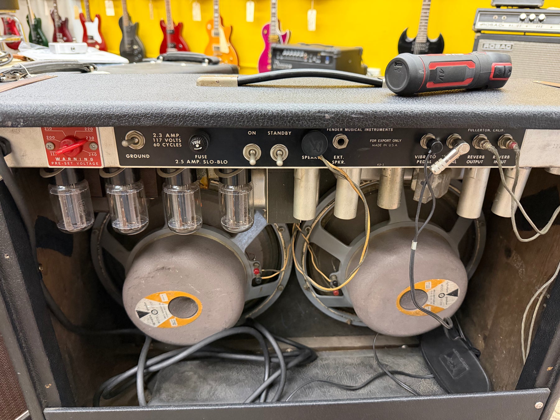 Rear view of a black amp, showing four glowing tubes, two large gray speakers, and the silver control panel with various inputs.