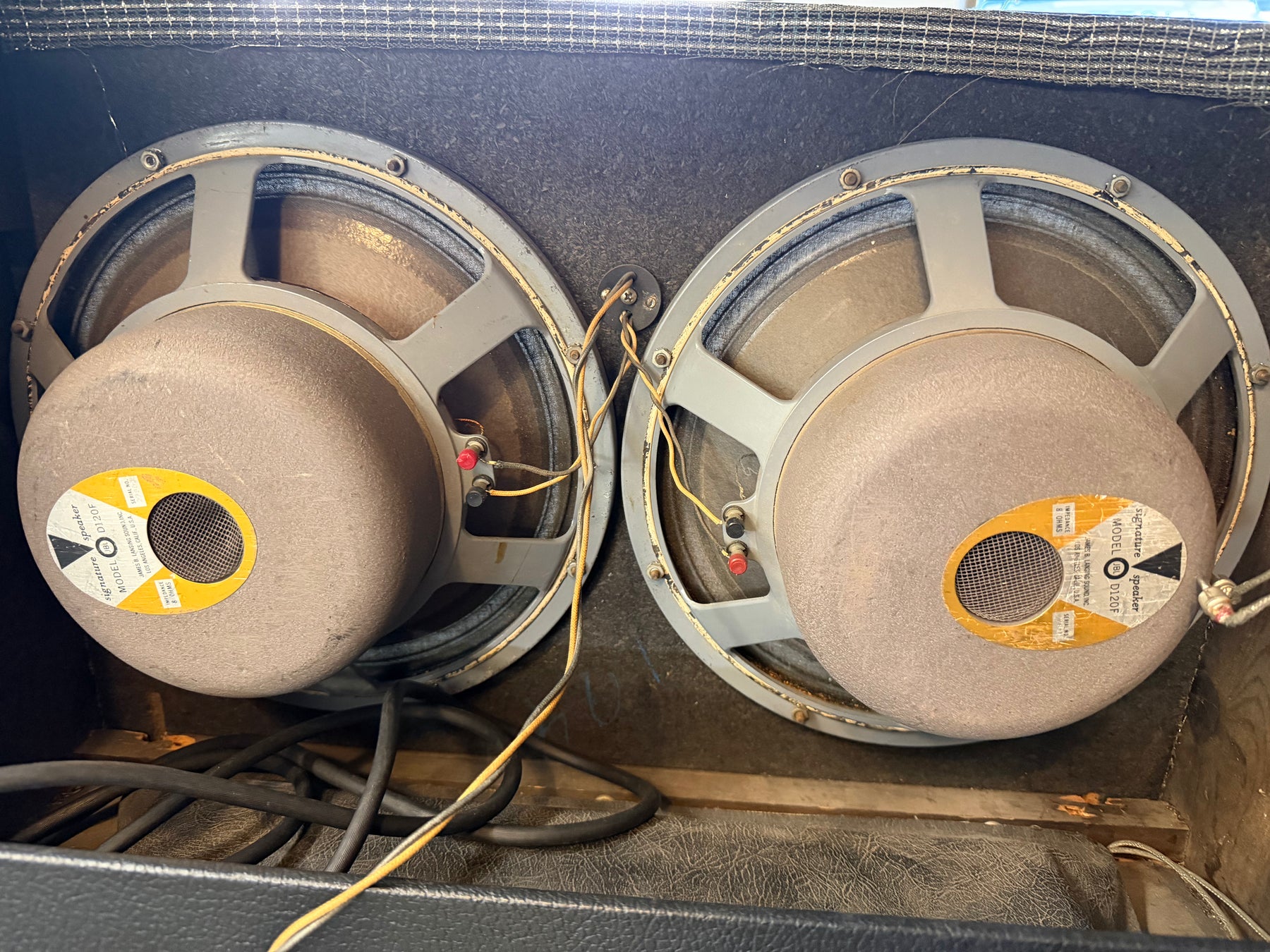 Two large, round, light gray speakers with tan cones and yellow labels are mounted inside a dark cabinet, showing wiring.
