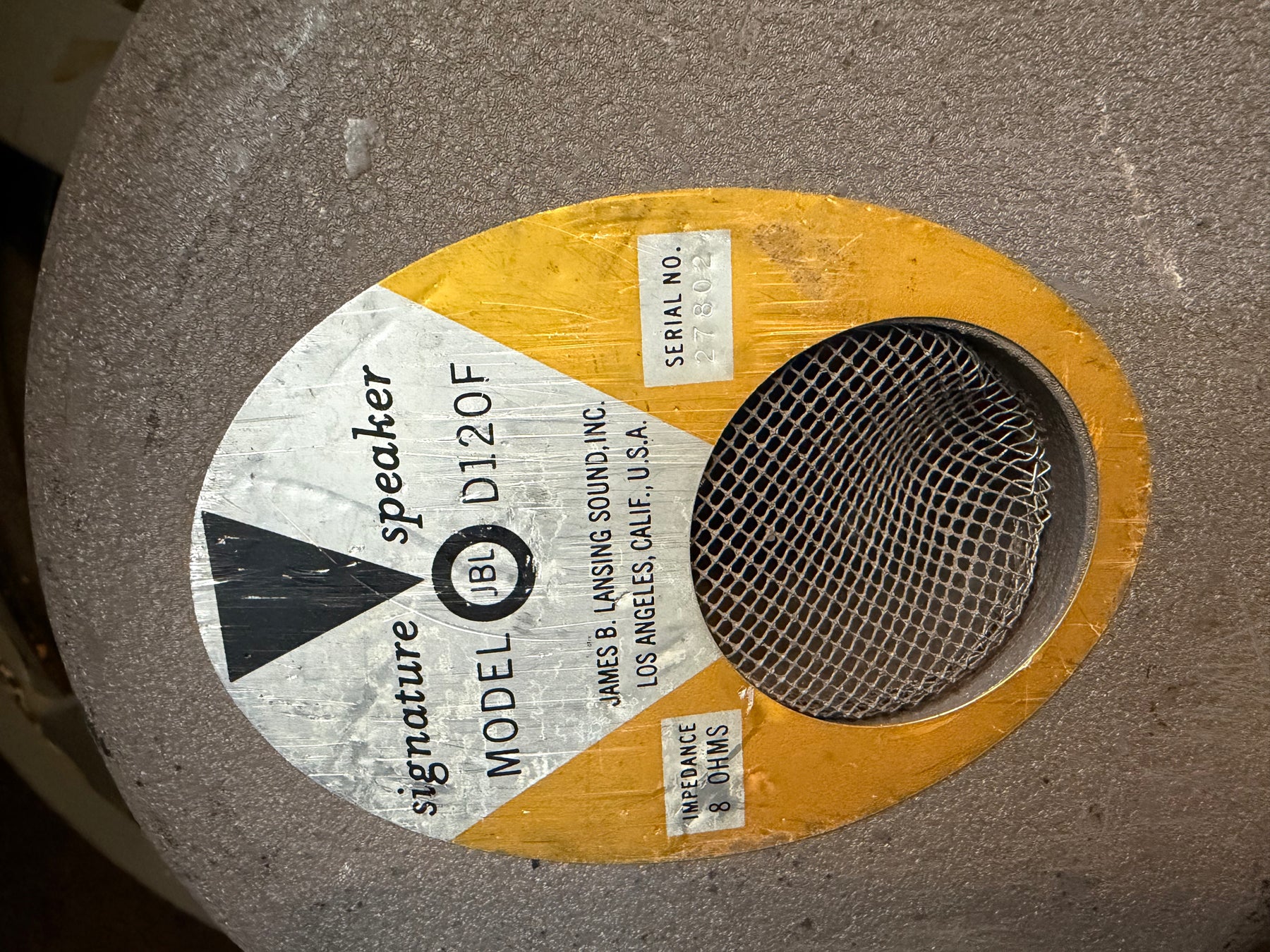 Close-up of a vintage JBL D120F speaker's gold and silver label, showing the mesh dust cap and some wear on the cone.