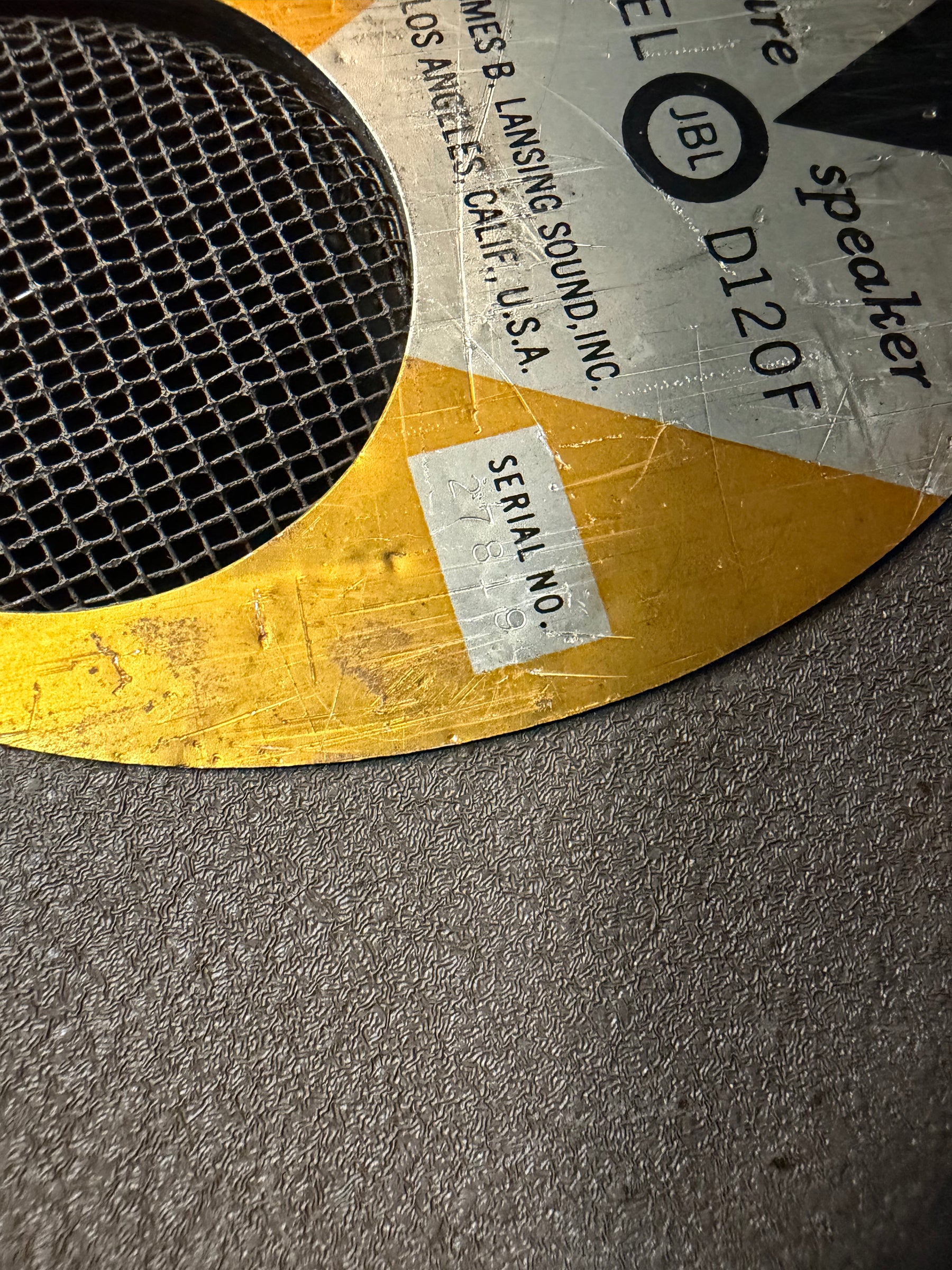 Close-up of a speaker's gold and silver frame with a metal mesh, showing visible scratches and a serial number sticker.