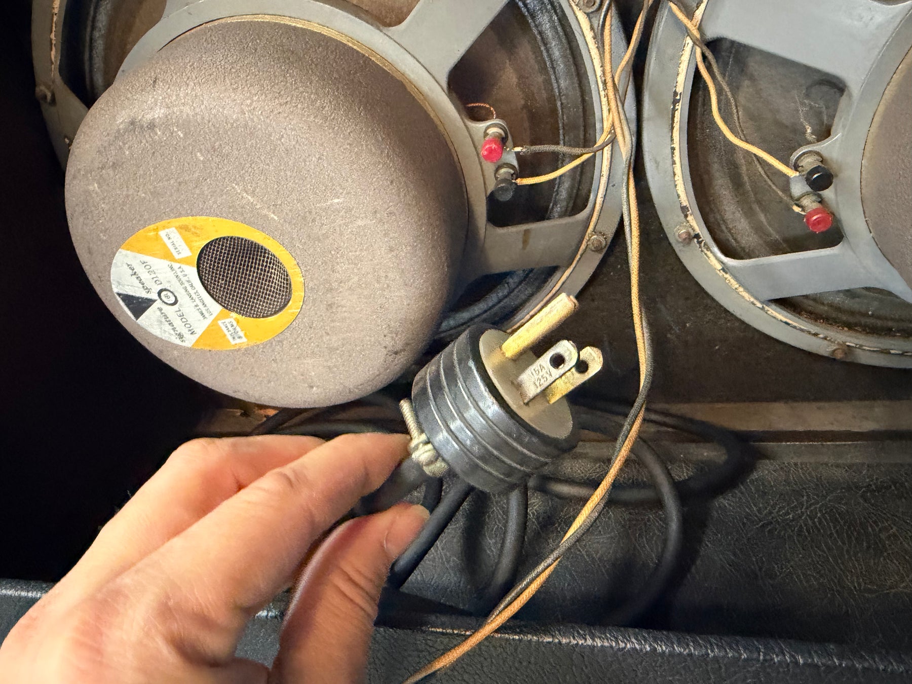 Close-up of the amp's two gray-framed speakers with exposed wiring, a hand holding a power plug, and a yellow label on the left speaker.