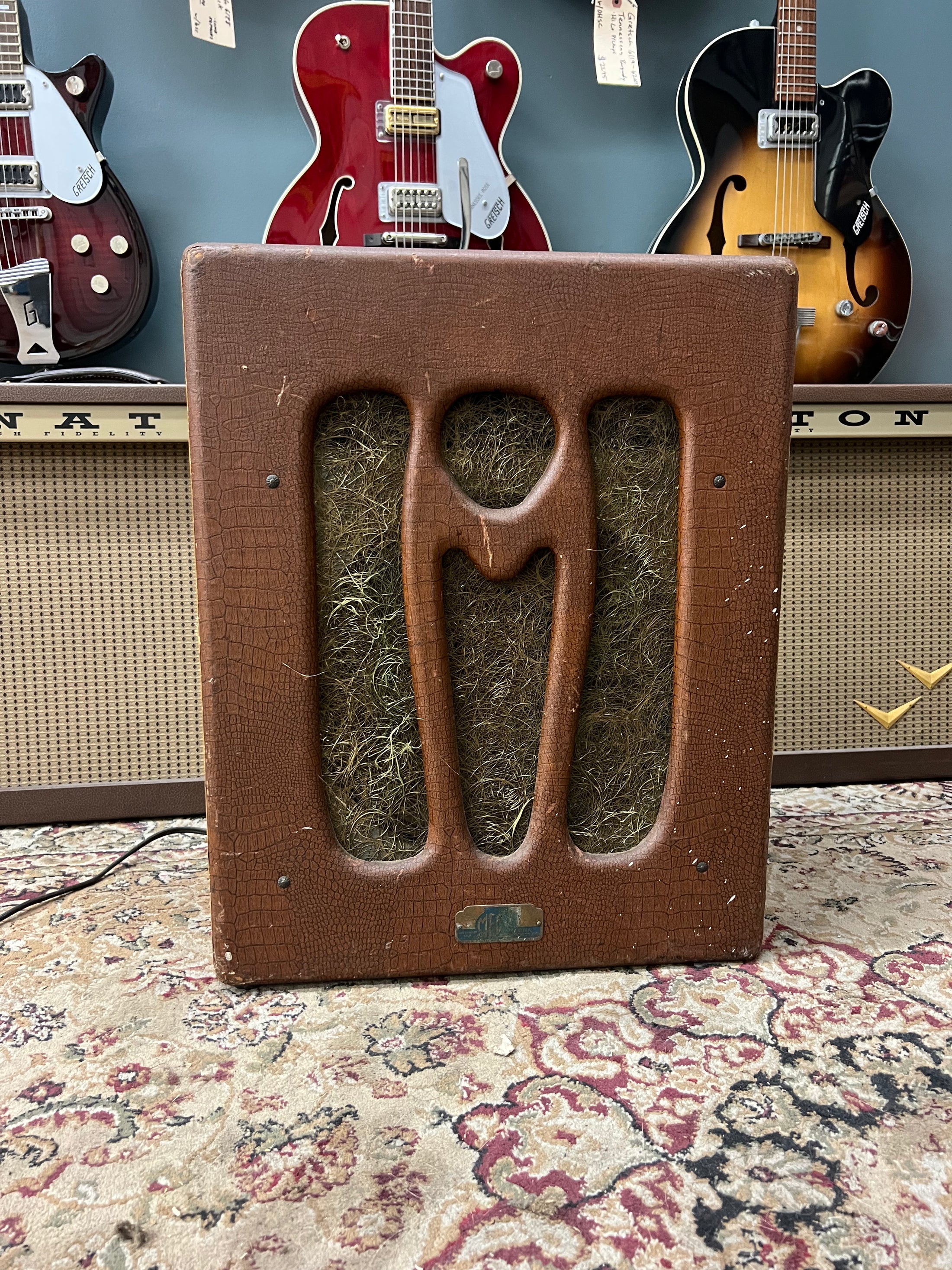 Front view of a brown alligator-textured combo amp with a stylized "M" cutout over a straw-colored grille cloth, showing wear.