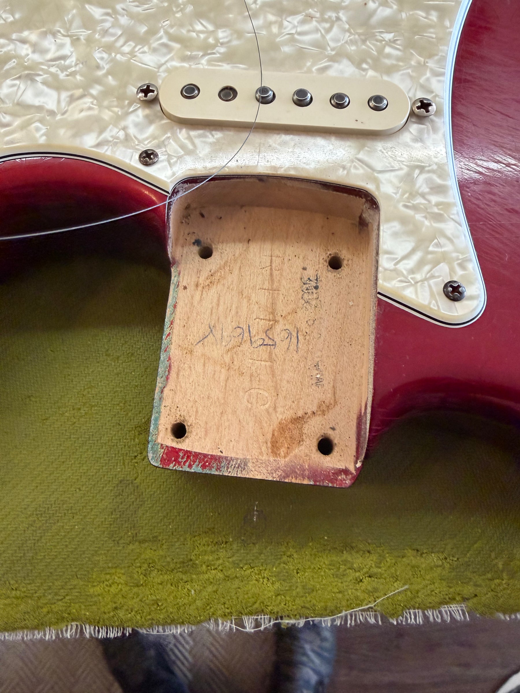 Close-up of the neck pocket, revealing raw wood with handwritten markings, surrounded by the red body and pearloid pickguard.