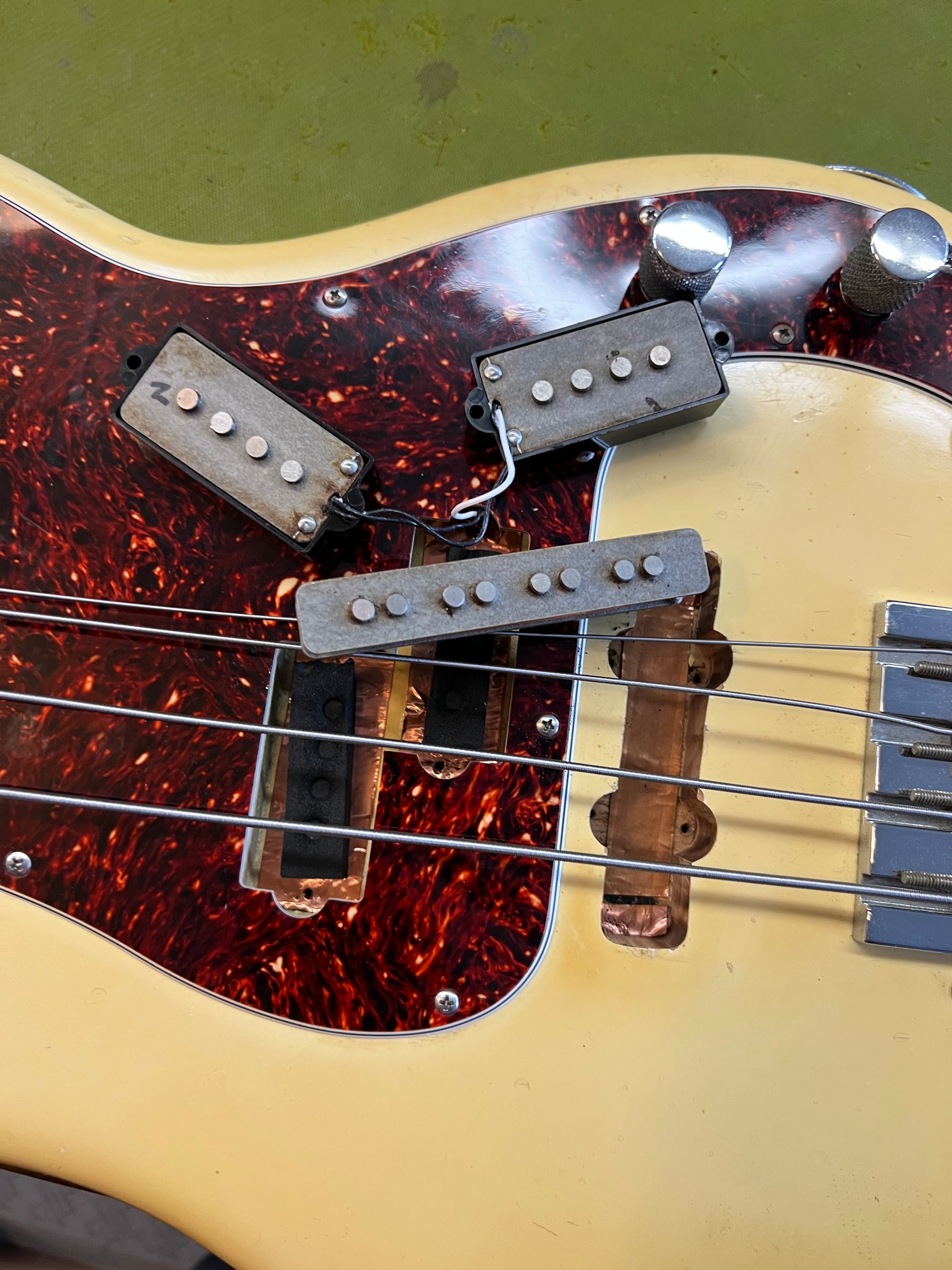 Close-up of a cream bass guitar with a tortoiseshell pickguard, showing three detached pickups and exposed wiring.