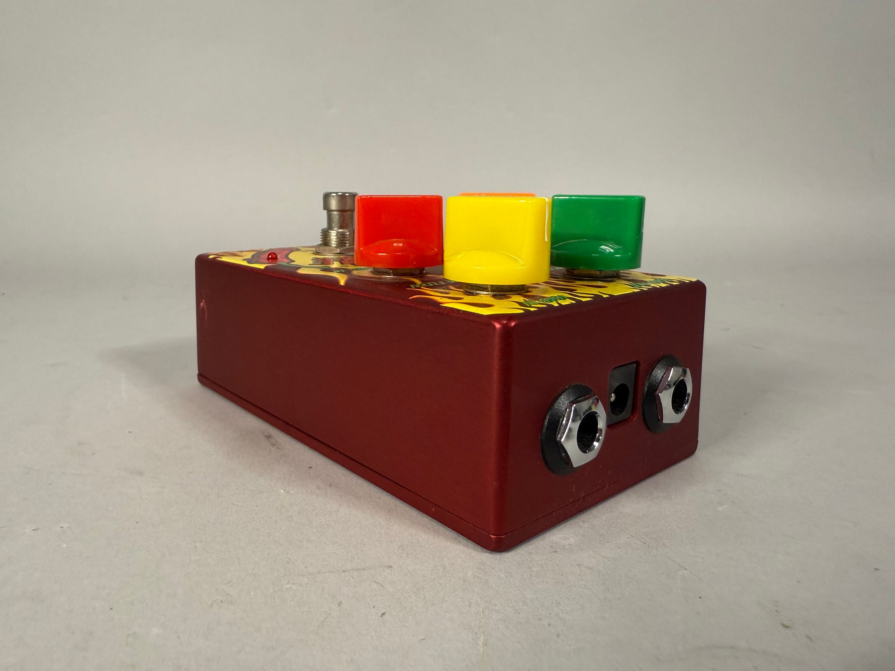 The red pedal is angled to show its right side with two input jacks, and the top with a sunflower pattern and three colorful knobs.