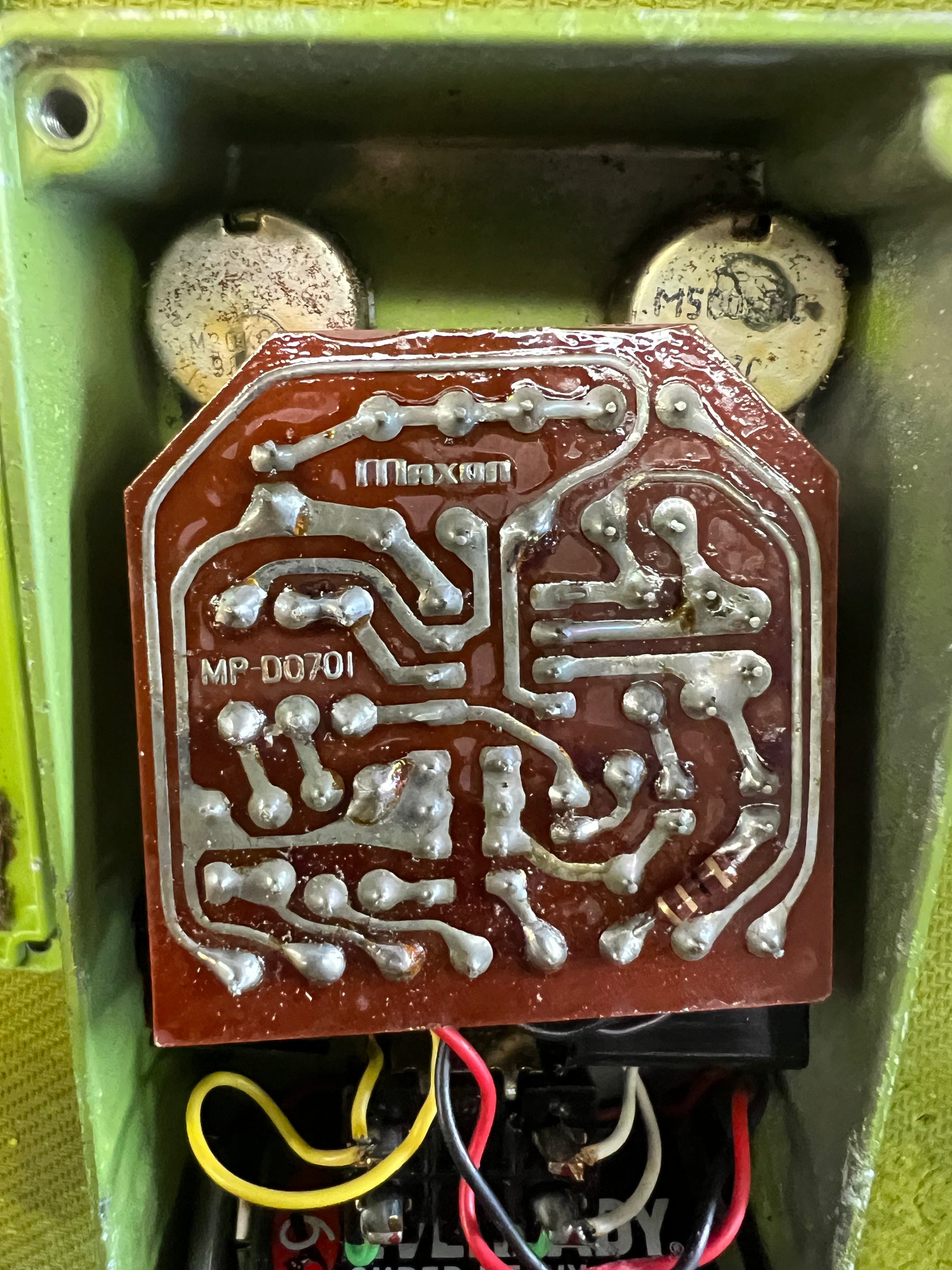 Close-up of the pedal's open chassis, revealing a brown circuit board with silver traces and visible wiring, set in a green enclosure.