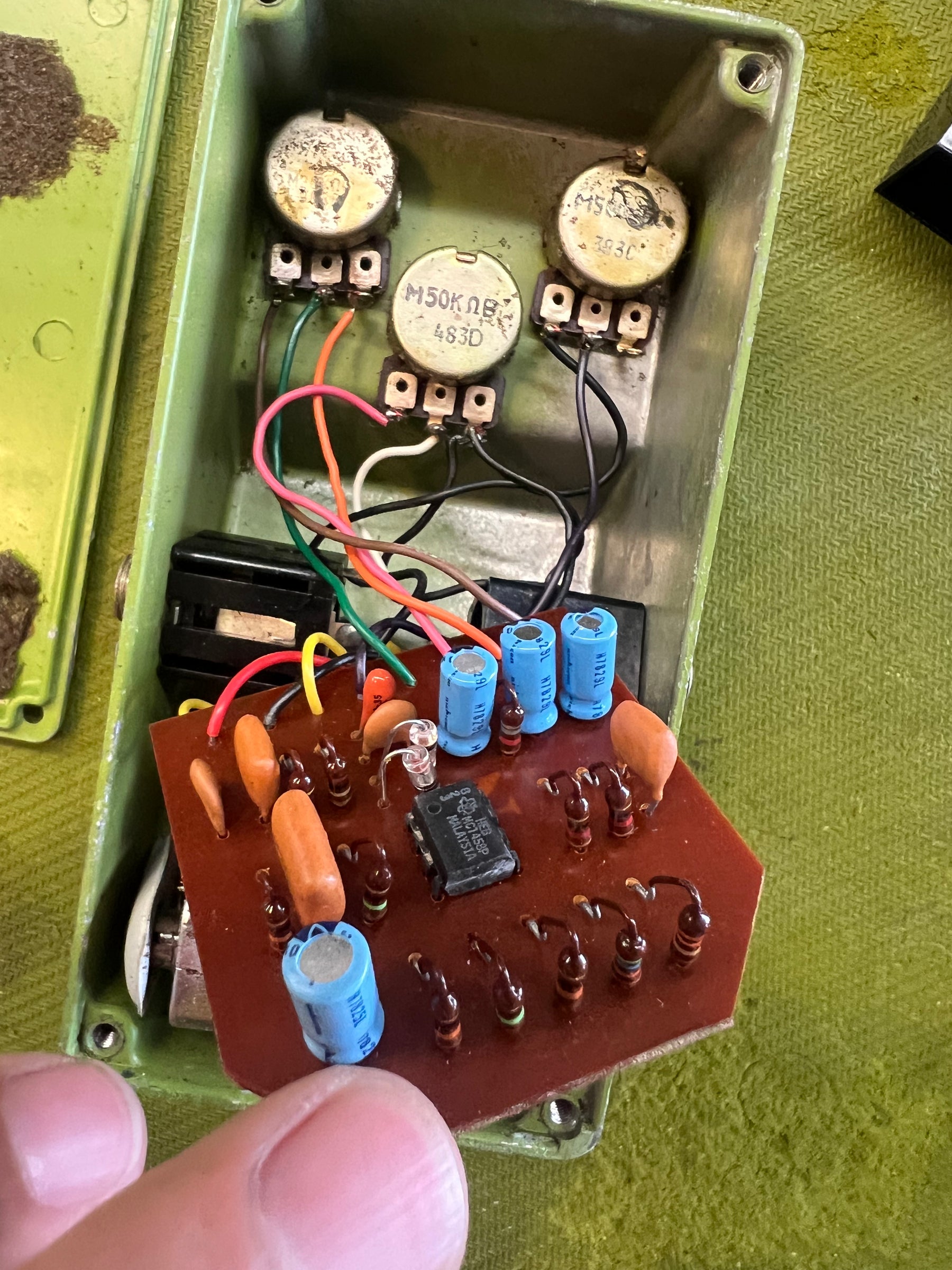 An overhead view of the open green pedal housing reveals the circuit board with blue capacitors, orange resistors, and three metal potentiometers.