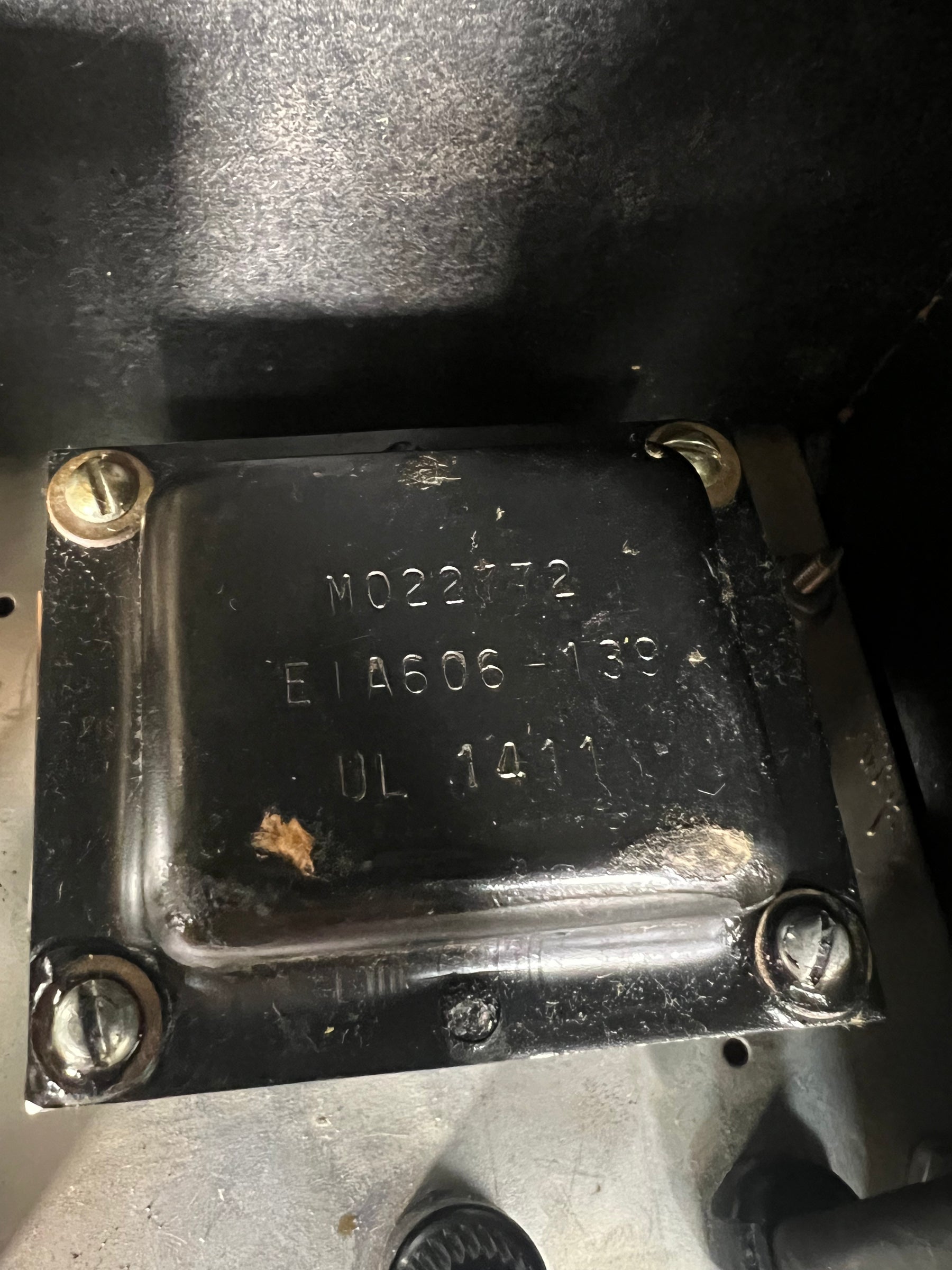 Close-up of a black metal transformer with visible numbers and letters, secured by four brass screws, showing some dust and wear.