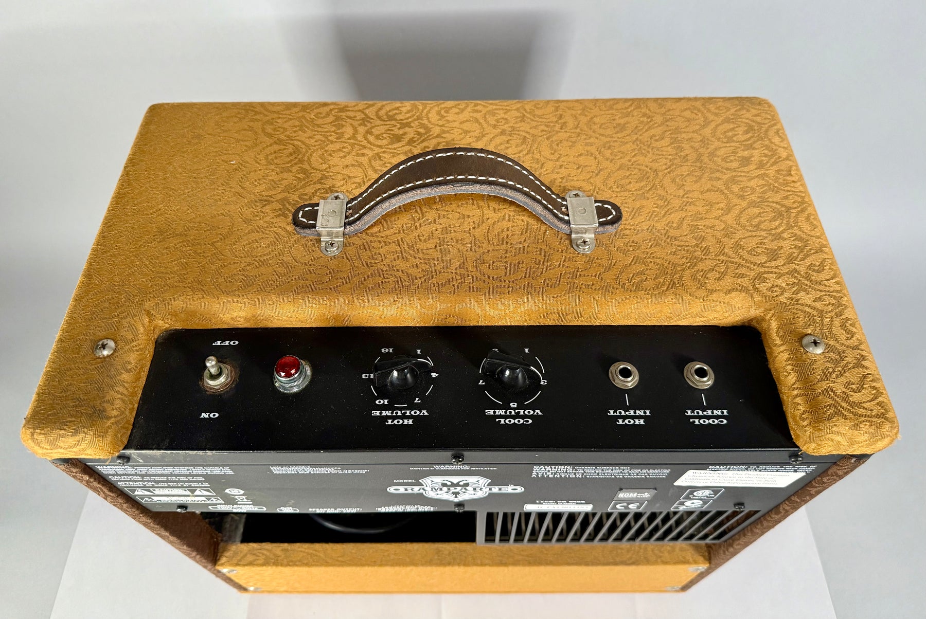 Top-down view of a gold patterned amp with a brown leather handle, showing the black control panel with two volume knobs and two input jacks.