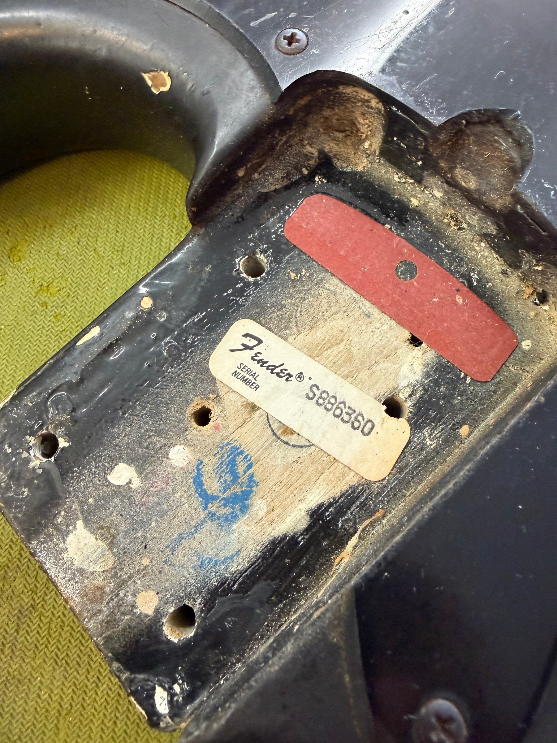 Close-up of the black bass body's neck pocket, showing exposed wood, a serial number sticker, and a red shim.
