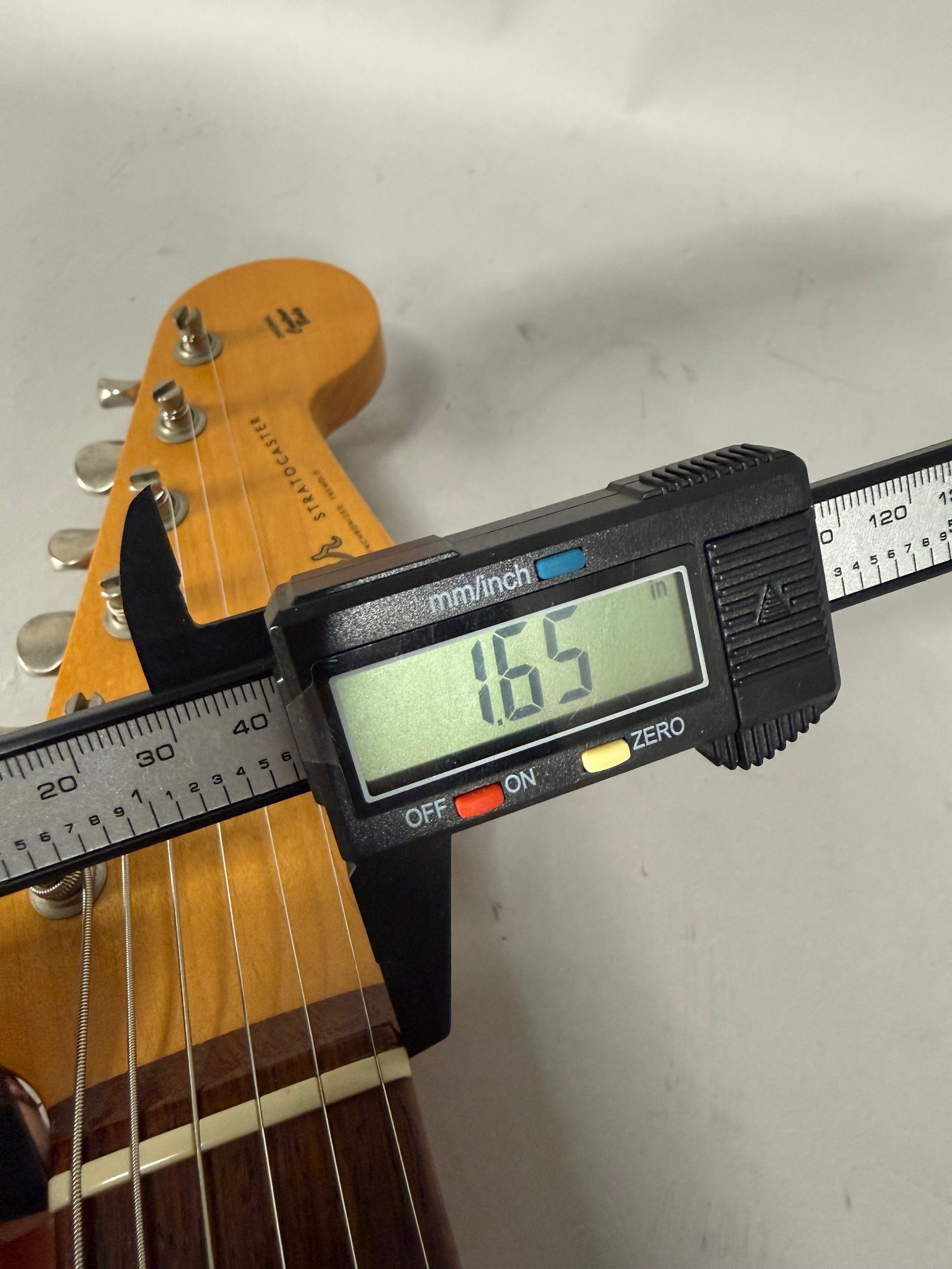 A digital caliper measures the guitar's nut width at 1.65 inches, with the headstock and tuning pegs visible in the background.