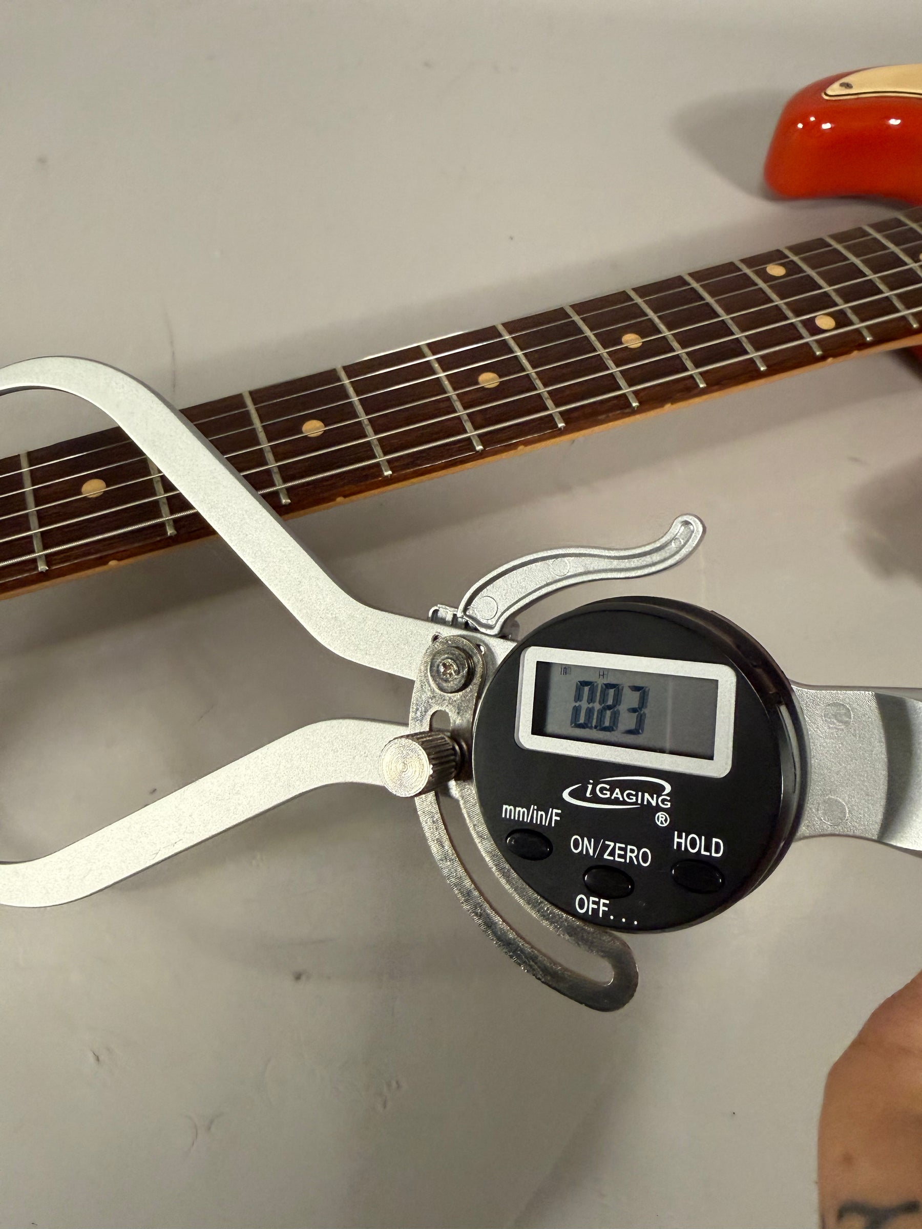 A digital caliper measures the guitar's dark fretboard, showing "0.83" on its screen, with a Fiesta Red body visible at the top right.