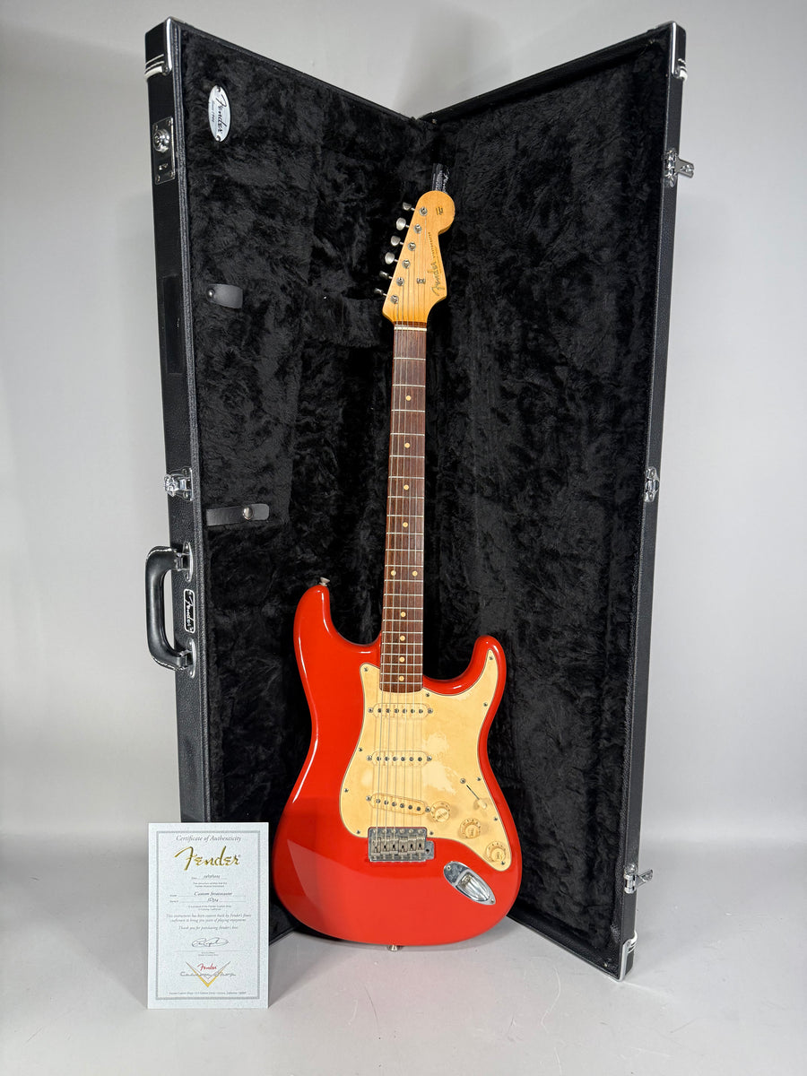 Full-length view of a glossy red Stratocaster with a cream pickguard, in an open black hard case, with a certificate of authenticity.
