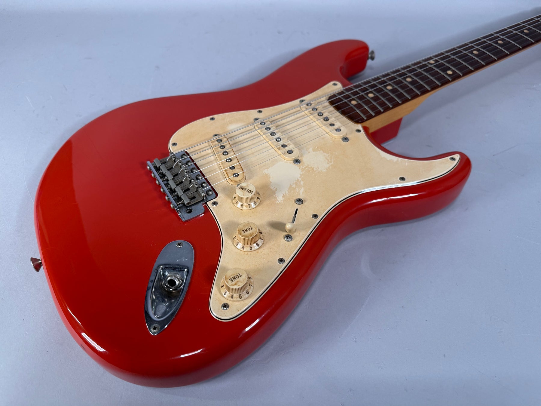Red electric guitar, angled view of the body and neck, showing a worn cream pickguard, three single-coil pickups, and chrome hardware.